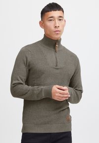 Hochkanziger olivgrüner Pullover mit strukturiertem Muster, gerippten Bündchen und Saum sowie einem Reißverschluss mit Leder-Ziehlasche. Logo-Patch an der Seite.