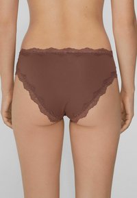 Femme portant de la lingerie marron avec des dentelles, vue de dos, sur un fond gris clair uni.