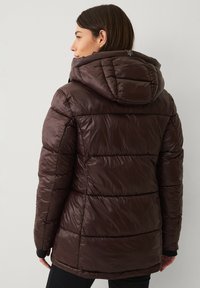 Kvinde iført en mørkebrun polstret jakke med hætte, set bagfra mod en ensfarvet lys baggrund.