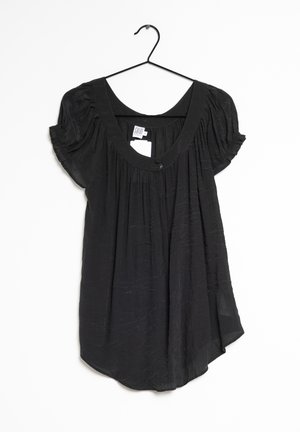 Blouse noire à manches courtes avec encolure dégagée et tissu froncé, suspendue sur un cintre noir sur fond blanc.