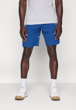 Niebieskie sportowe spodenki z gładkiej tkaniny, z bocznymi kieszeniami i kontrastowymi białymi szwami, noszone z szarą koszulką i białymi tenisówkami.