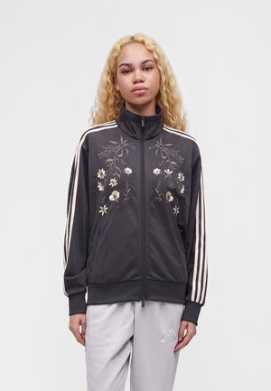 Jeune personne aux longs cheveux blonds bouclés porte une veste noire zippée avec broderies florales et manches rayées, associée à un pantalon gris clair.