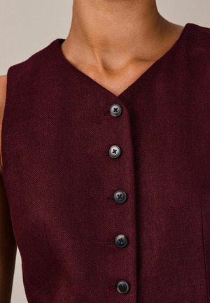 Ærmeløs bordeaux knaplukket beklædningsgenstand med V-udskæring båret af en person, der viser stoffets tekstur og fem sorte knapper ned foran.