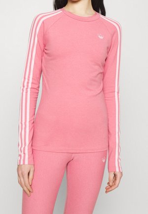 Personne portant un haut à manches longues rose et un legging assorti avec des détails blancs à trois bandes Adidas sur les manches et les côtés du pantalon.