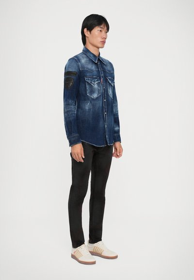 Junger Mann in einem verblassten blauen Jeanshemd mit Brusttaschen, schwarzen Hosen und weißen Sneakers mit beigen Streifen vor einem einfarbigen Hintergrund.