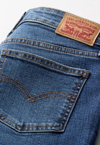 Jean en denim de teinte moyenne, avec des coutures orange contrastantes et une étiquette en cuir marron à la taille ornée de motifs rouges.
