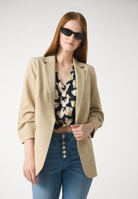 Beige Blazer mit strukturierten Revers, kombiniert mit einem marineblauen, blumigen Crop-Top. Das Model trägt hellblaue, hoch geschnittene Jeans mit Knopfdetails.