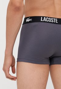 Szürke boxeralsó fekete elasztikus derékkal, amelyen fehér "Lacoste" logó található. Puha anyag, szoros illeszkedés és minimális varrás jellemzi.
