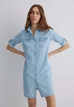 Light blue denim shirt dress with button-down front, two chest pockets, and rolled sleeves. Smooth texture and knee-length design.