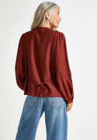 Rustfarvet bluse med plisseret tekstur, pufærmer og snøre detaljer i taljen, parret med lyseblå wide-leg jeans.