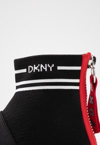 Černá kotníková bota s žebrovaným límcem, bílými proužkovými akcenty a detailem červeného zipu. Na límci je logo DKNY.