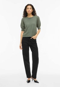 Vrouw met krullend haar, gekleed in een groen topje met pofmouwen, zwarte hoge taillebroek en zwarte pumps met een puntige neus, staand tegen een witte achtergrond.