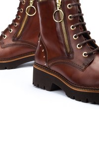 Botas de tobillo de cuero marrón con cremalleras doradas, cordones redondos, acentos de tachuelas, suela gruesa y con un dibujo resistente para mayor tracción.