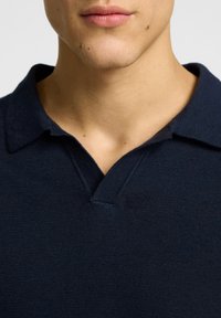 Navy blue polo shirt with short sleeves, a V-shaped collar, and a textured knit fabric. Stitching detail visible along the collar.