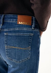 Tmavě modré džínové kalhoty se hnědou koženou nášivkou s logem "MEXX" ve zlaté barvě. Obsahují zadní kapsy a dekorační švy.