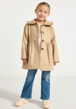 Ragazza giovane che sorride, indossa un cappotto beige con bottoni e colletto arricciato, jeans blu e scarpe da ginnastica beige, in piedi davanti a uno sfondo chiaro.