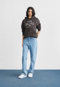 Καφετί φούτερ με κουκούλα με την εκτύπωση «New Balance Athletics», συνδυασμένο με ανοιχτό μπλε τζιν με ρίγες και λευκά αθλητικά παπούτσια σε φόντο μπλε χαλιού.