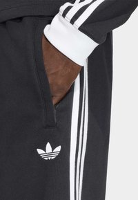 Černé sportovní tepláky s bílými třemi proužky, elastickým pasem a bočními kapsami. Na noze je malý bílý logo Adidas.