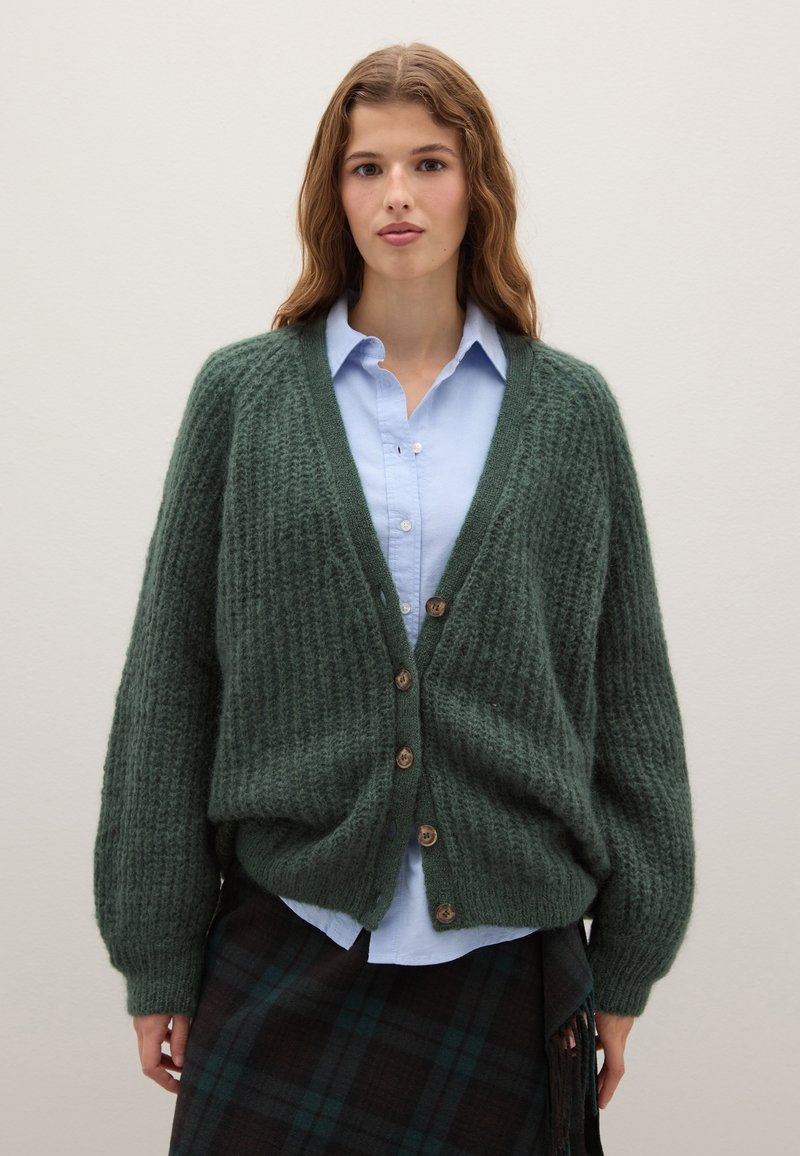 Cardigan verde a maglia con vestibilità ampia, texture a coste e bottoni in legno. Indossato sopra una camicia azzurro chiaro e una gonna a quadri.