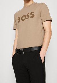 Beige bomulls-t-shirt med stort brunt "BOSS"-tryck, kombinerad med svart bälte och texturerade svarta byxor med rak skärning.