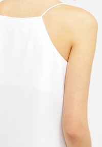 Top bianco senza maniche con spalline sottili, tessuto liscio e una leggera brillantezza, che evidenzia i dettagli del collo e delle spalle.