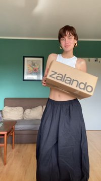Kartonová krabice s logem "zalando" v tučném černém písmu, držená osobou v bílém vršku a dlouhé černé sukni, vevnitř.