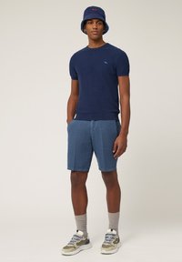 Marine gebreide T-shirt trui, blauwe linnen shorts, grijze sokken en sneakers. De bucket hat heeft een vergelijkbaar kleurenschema met rode accenten.