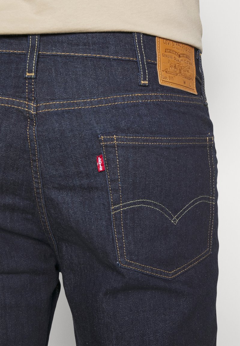 Jean en denim noir avec des coutures jaunes, comportant un patch en cuir et une petite étiquette rouge sur la poche arrière. Design slim fit classique.