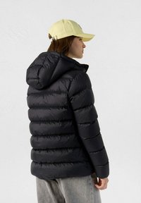 Kvinde iført en sort puffjakke med hætte og lysegul baseballkasket, der står sidelæns mod en ensfarvet grå baggrund.