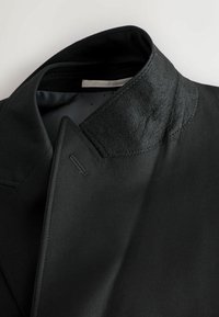 Jachetă neagră cu un guler structurat, având o combinație de țesătură netedă și lână texturată. Detalii de cusătură notabile și un design elegant.