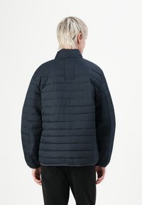 Sötétkék steppelt kabát magas nyakkal, vízszintesen steppelt panelekkel és lazán illeszkedő fazonnal. Texturált anyag, rejtett varrási részletekkel.