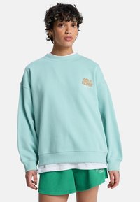 Φωτεινό μέντα πράσινο φούτερ κατασκευασμένο από απαλό υλικό, με μεγάλο πορτοκαλί γραφικό στο αριστερό στήθος και χαλαρή εφαρμογή.