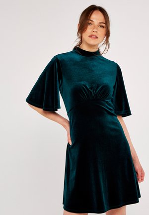 Vestido de terciopelo verde azulado con mangas cortas y acampanadas, un corsé entallado y detalle de cintura fruncida. Textura suave y corte a la altura de la rodilla.