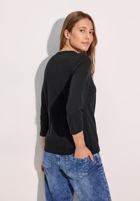 Schwarzes, langärmliges Damenoberteil mit rundem Ausschnitt, aus weichem Stoff; kombiniert mit hellblauen, jeansähnlichen Hosen mit Gesäßtaschen.