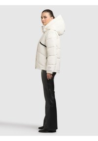 Biała kurtka puchowa z dużym kapturem, zapinana na zamek z przodu, z czarną kieszenią na zamek. W połączeniu z czarnymi skórzanymi spodniami i butami.