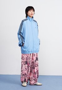 Chaqueta de chándal azul claro con rayas negras, combinada con pantalones de pierna ancha de estampado floral rosa. La modelo está de pie sobre un suelo azul.