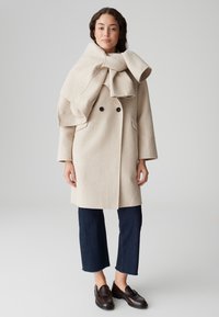Beige wollen double-breasted jas met een grote gevouwen kraag, gecombineerd met cropped donkere spijkerbroeken en bruine loafers. Zachte textuur door het hele kledingstuk.