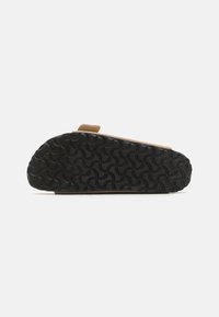 Sandale en suede marron avec une semelle en caoutchouc noir texturé, présentant un motif ondulé pour une meilleure adhérence et une seule sangle sur le dessus pour un bon maintien.