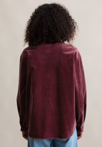 Bordeauxrode ribfluwelen blouse met een losse pasvorm, lange mouwen, geribbelde textuur en knoopmanchetten, van achteren getoond tegen een neutrale achtergrond.