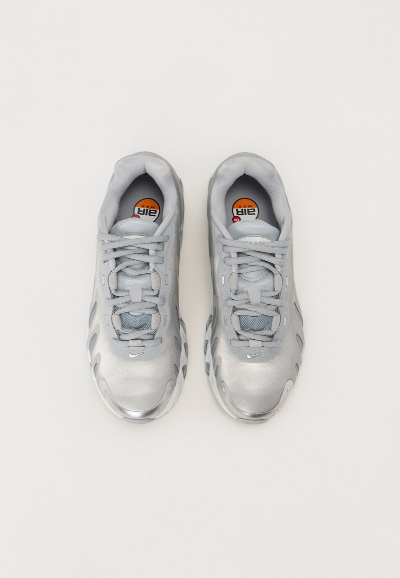 Zapatillas deportivas plateadas con una parte superior suave, acentos de malla y suelas texturizadas. Presentan una puntera redondeada y cordones grises claros.