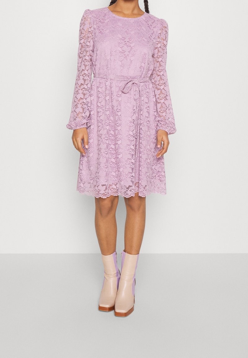 Femme portant une robe en dentelle lavande jusqu’aux genoux avec manches longues et ceinture à nouer, associée à des bottines bicolores beige et lavande.