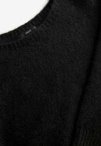 Pull en maille noire avec un col rond, un tissu texturé et une étiquette "NEXT" au niveau du col. Apparence chaude et douce.