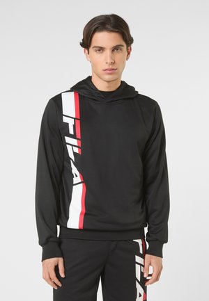 Junger Mann mit kurzen dunklen Haaren, der einen schwarzen FILA-Kapuzenpullover und passende Hosen mit roten und weißen vertikalen Logostreifen trägt.