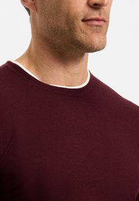 Gros plan d'un homme portant un pull bordeaux à col rond avec un sous-vêtement blanc visible au niveau du col, montré du cou à la poitrine.