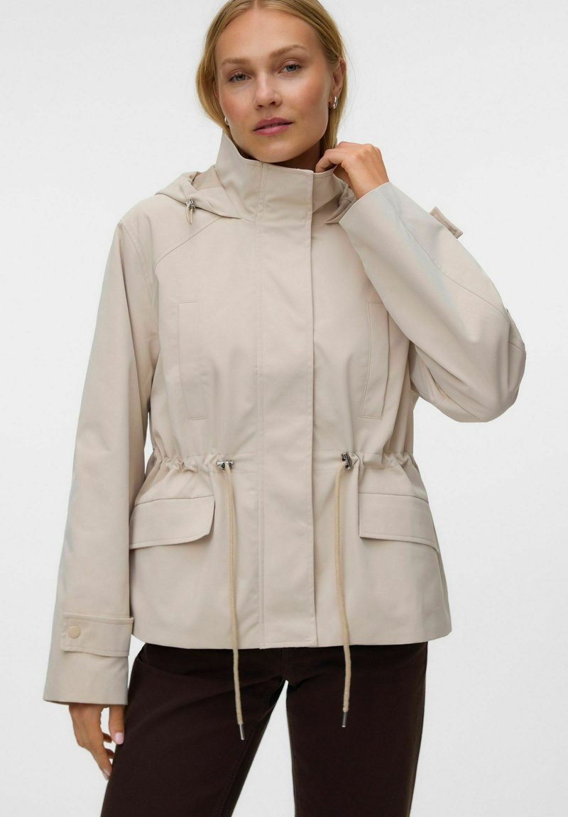 Femme portant une veste beige à capuche avec cordons à la taille et poches à rabat, associée à un pantalon foncé, posant devant un fond uni.