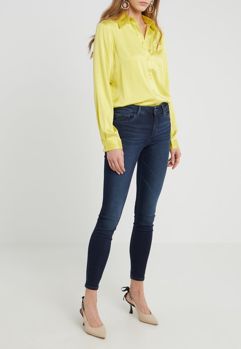 Žena oblečená v žluté saténové blůze, tmavých úzkých džínech, béžových podpatcích s černými členkovými pásky a velkých kruhových náušnicích stojí na bílém pozadí.