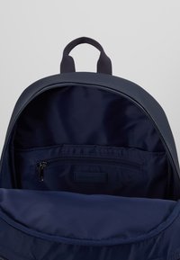 Tummansininen reppu, jossa on avoin pääosasto, sisempi vetoketjullinen tasku ja kangaslenkki ylhäällä.