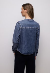 Femme aux cheveux longs châtain clair portant une chemise en jean bleu délavé et un pantalon foncé, debout devant un fond gris clair uni.