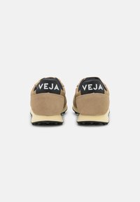 Beige semsket sko med svarte detaljer, med hæl som er hevet, hvit logo bak og teksturert svart gummisåle.