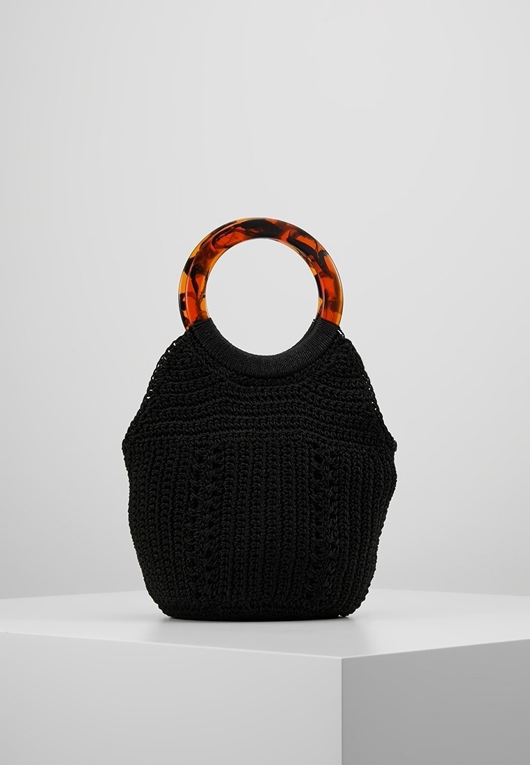 Zwart gebreide handtas met ronde handvatten van kunststof met schildpadmotief, tentoongesteld op een wit voetstuk tegen een grijze achtergrond.